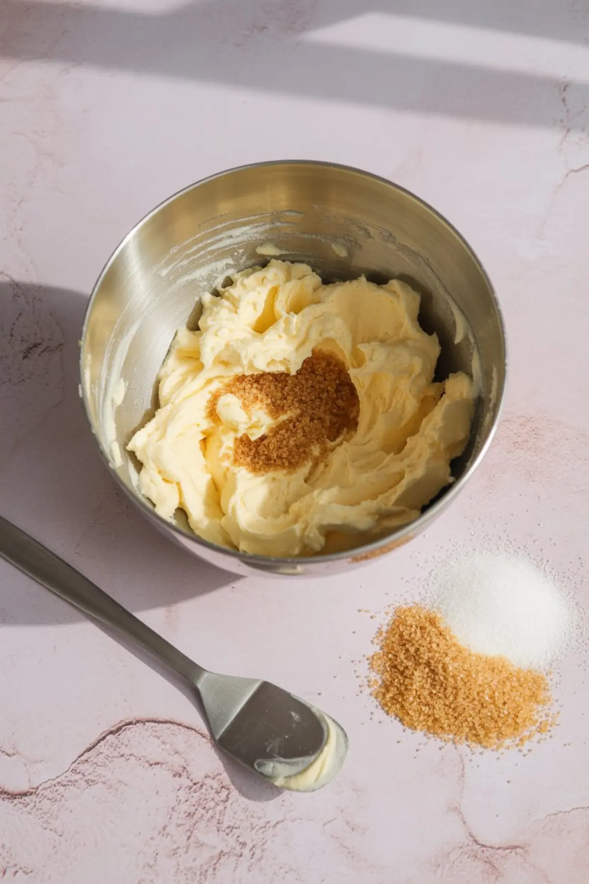 Metal mixing bowl filled with creamed butter and brown sugar, surrounded by scattered white and brown sugar piles, with a spatula resting beside it.
