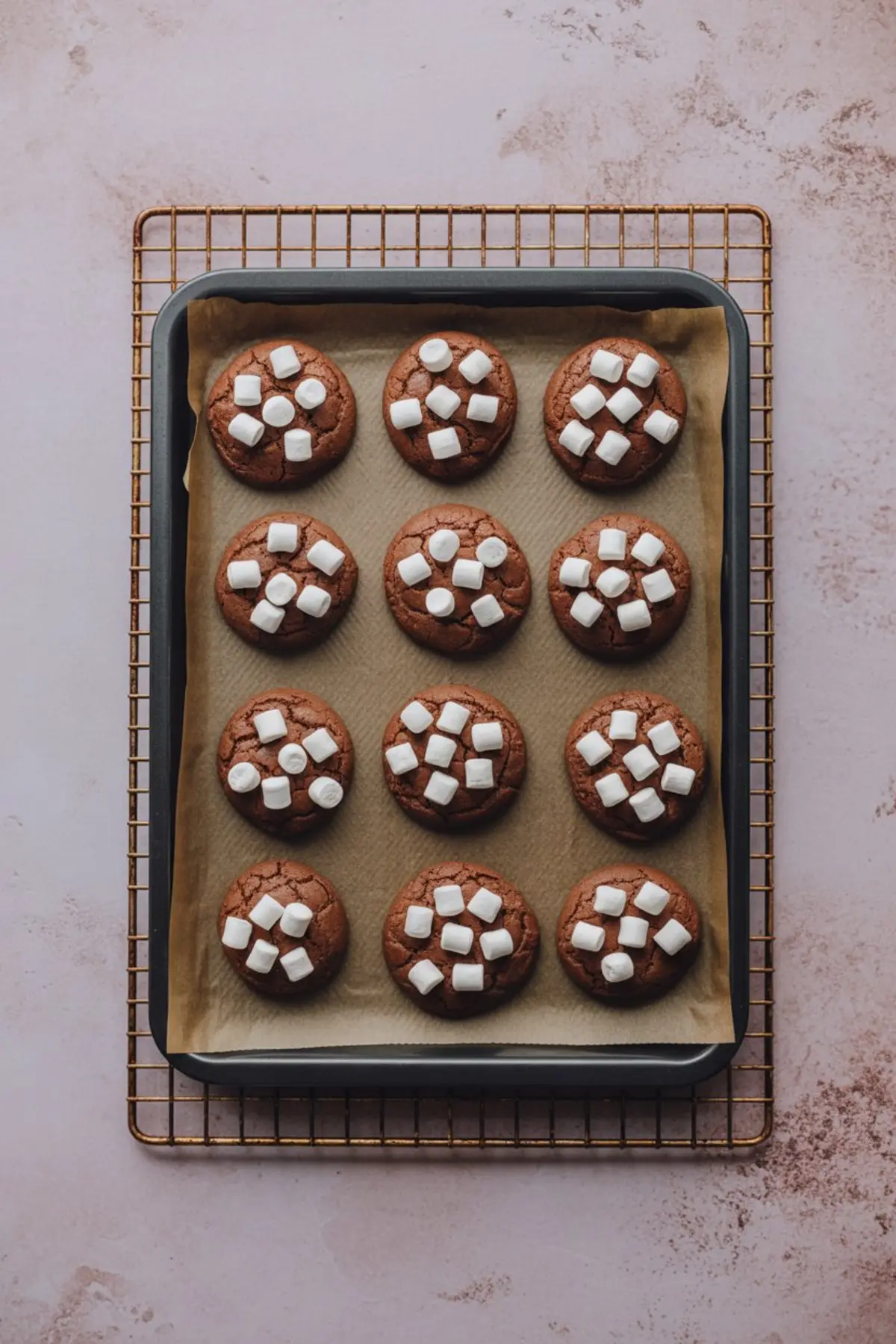 Tray of freshly baked chocolate cookies topped with mini marshmallows on parchment paper, resting on a wire cooling rack over a soft pink surface.
