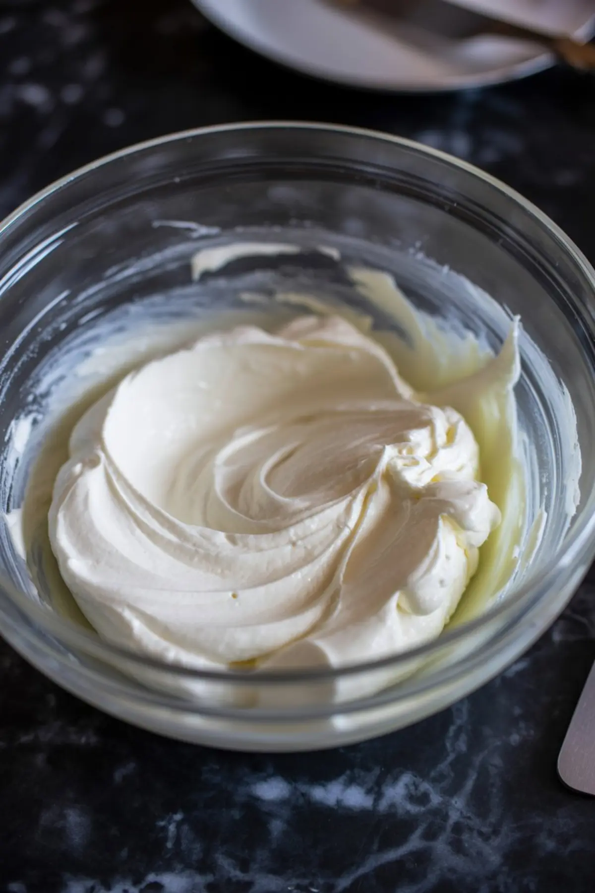 Glass bowl filled with whipped cream cheese mixture, showing light and fluffy texture for the cheesecake filling preparation on a dark counter.
