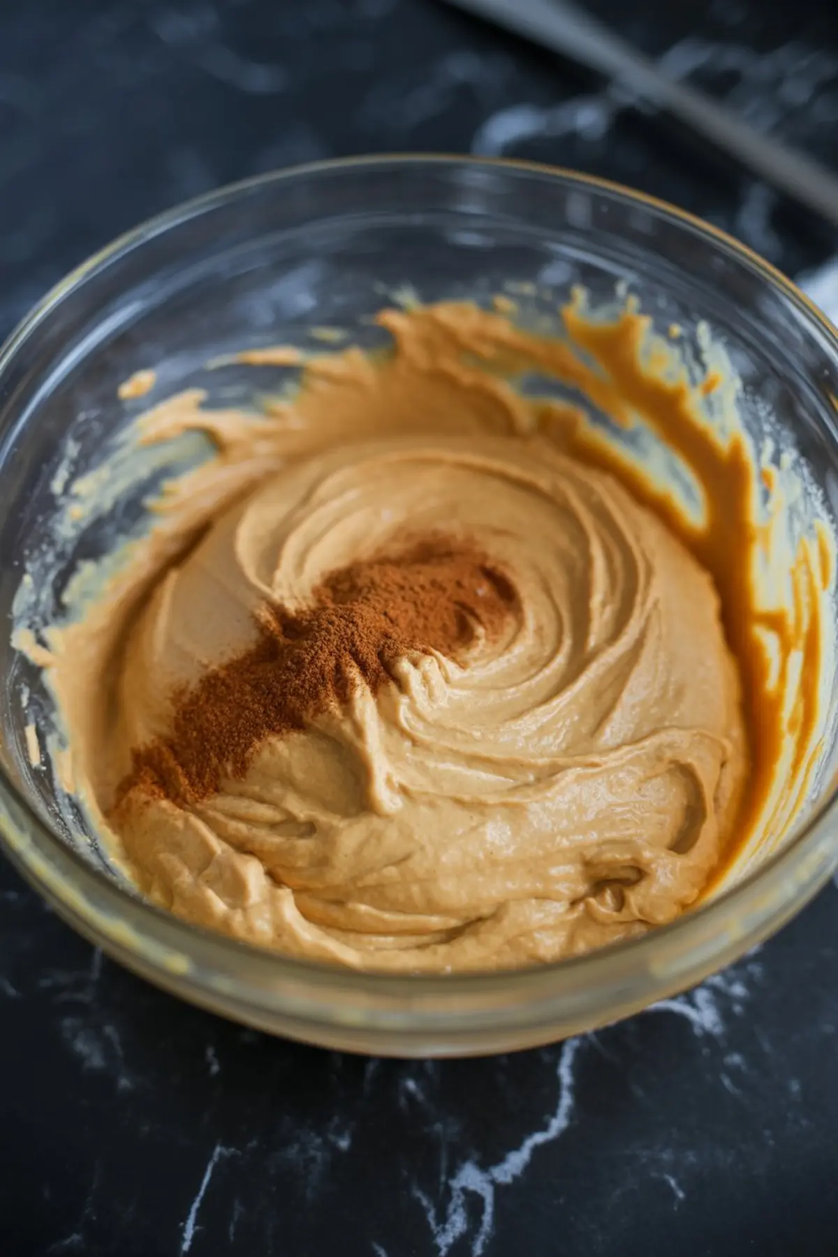 Bowl of creamy pumpkin cheesecake batter swirled with cinnamon, showing smooth texture and rich orange color on a dark marble background.
