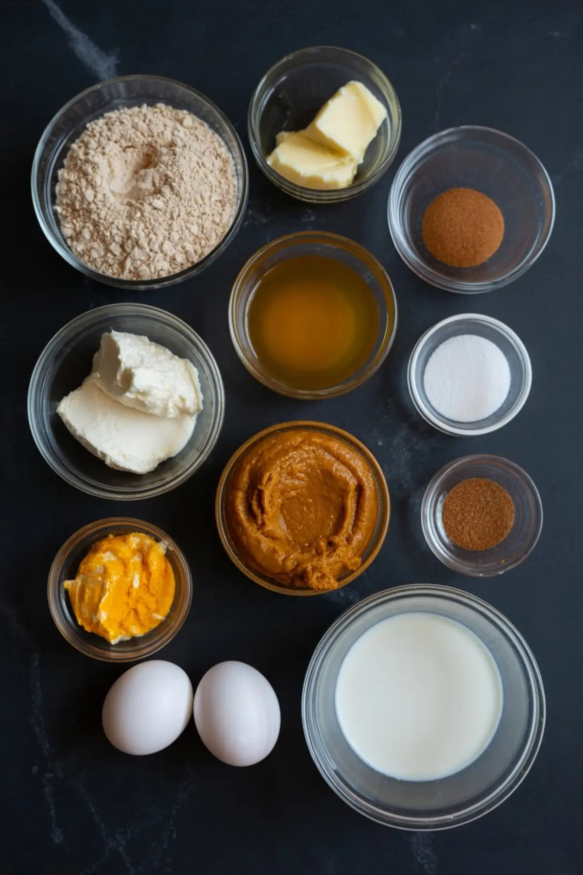 Top-down view of keto pumpkin cheesecake ingredients arranged in glass bowls, including almond flour, butter, eggs, cream cheese, pumpkin puree, sweetener, vanilla extract, and seasonal spices like cinnamon and nutmeg.
