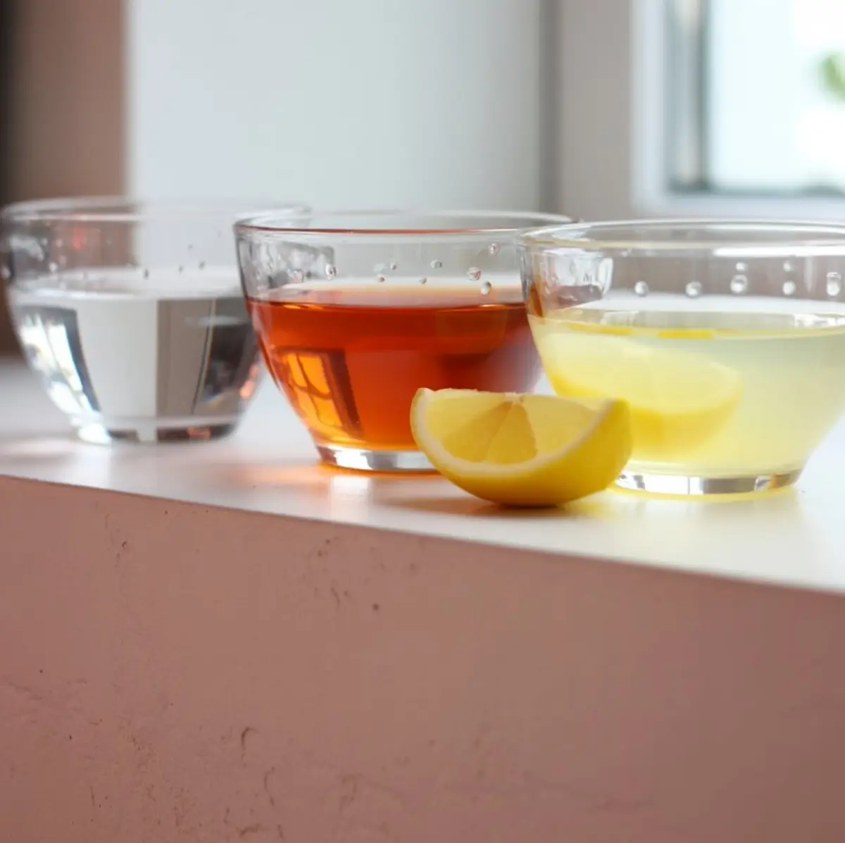 Three clear glass bowls filled with cocktail ingredients — sugar, amber-colored simple syrup, and pale yellow lemon juice — arranged on a white surface with a lemon wedge in front, ideal for homemade lemon drop martini preparation.
