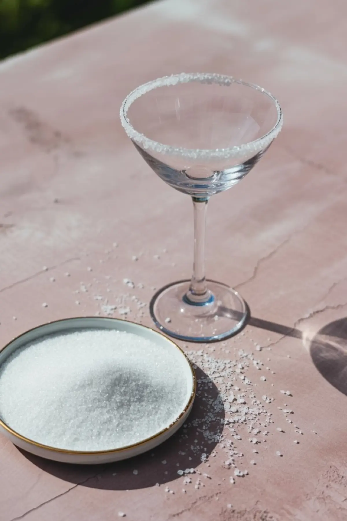 Empty martini glass with a sugared rim standing on a pink countertop, next to a shallow dish filled with white granulated sugar and scattered sugar crystals.
