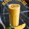 Tall glass of mango smoothie topped with swirl of syrup and cardamom seeds, styled with mango slice and mint leaves on a black marble countertop under the curved text “Mango Smoothie”.