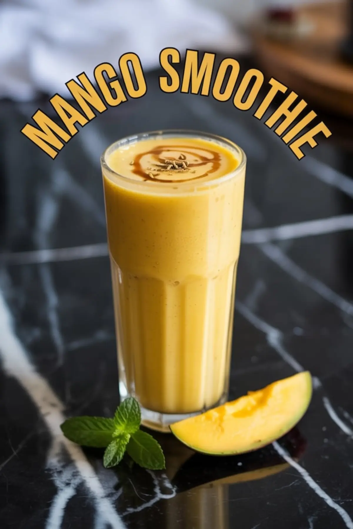 Tall glass of mango smoothie topped with swirl of syrup and cardamom seeds, styled with mango slice and mint leaves on a black marble countertop under the curved text “Mango Smoothie”.