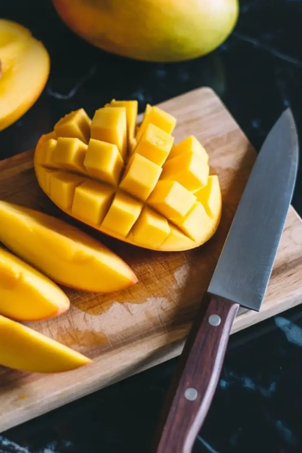 Cutting board with sliced and diced ripe mango, a halved mango with seed, and a whole mango, placed next to a kitchen knife on a dark stone surface.
