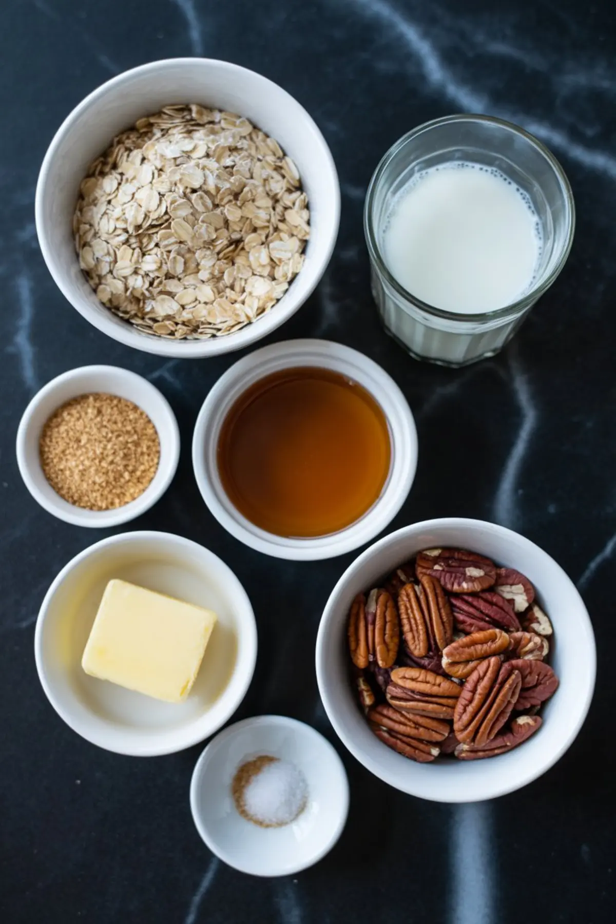 Flat lay of oatmeal ingredients including rolled oats, milk, maple syrup, brown sugar, butter, pecans, and salt arranged in white bowls on a black marble countertop.