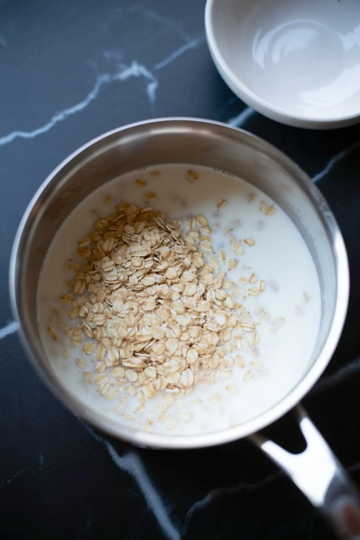 Rolled oats poured into a saucepan of milk, ready to cook for oatmeal on a dark marble surface.