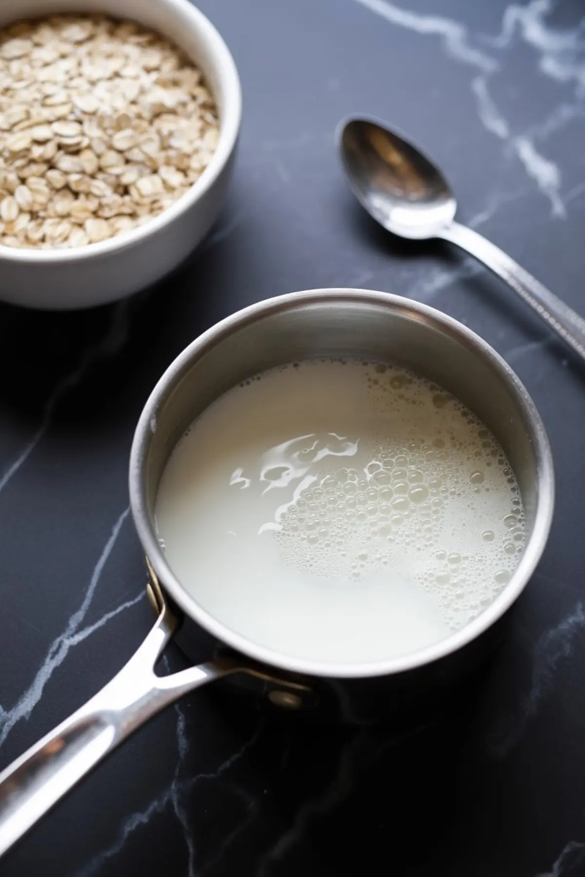 Stainless steel saucepan filled with milk on a black marble counter, with a spoon and a white bowl of rolled oats nearby.