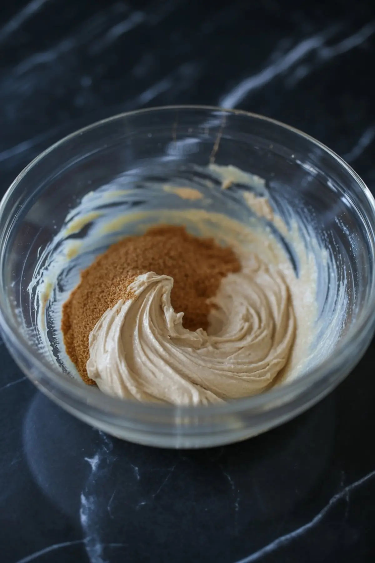 Cream cheese mixture partially blended with brown sugar in a glass bowl, showing soft whipped texture on a black marble surface.