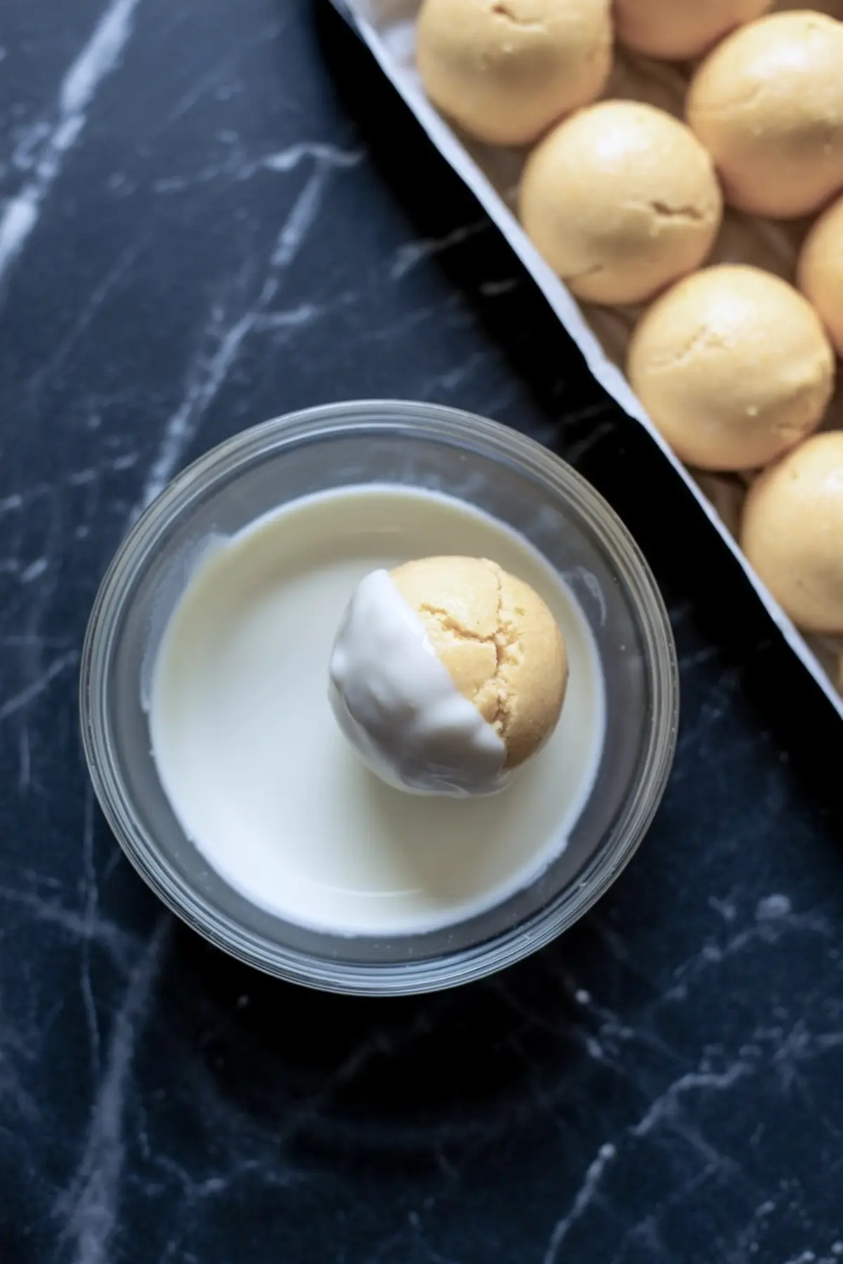 Round pumpkin cheesecake ball being dipped into a bowl of melted white chocolate with more cheesecake balls on a tray in the background.