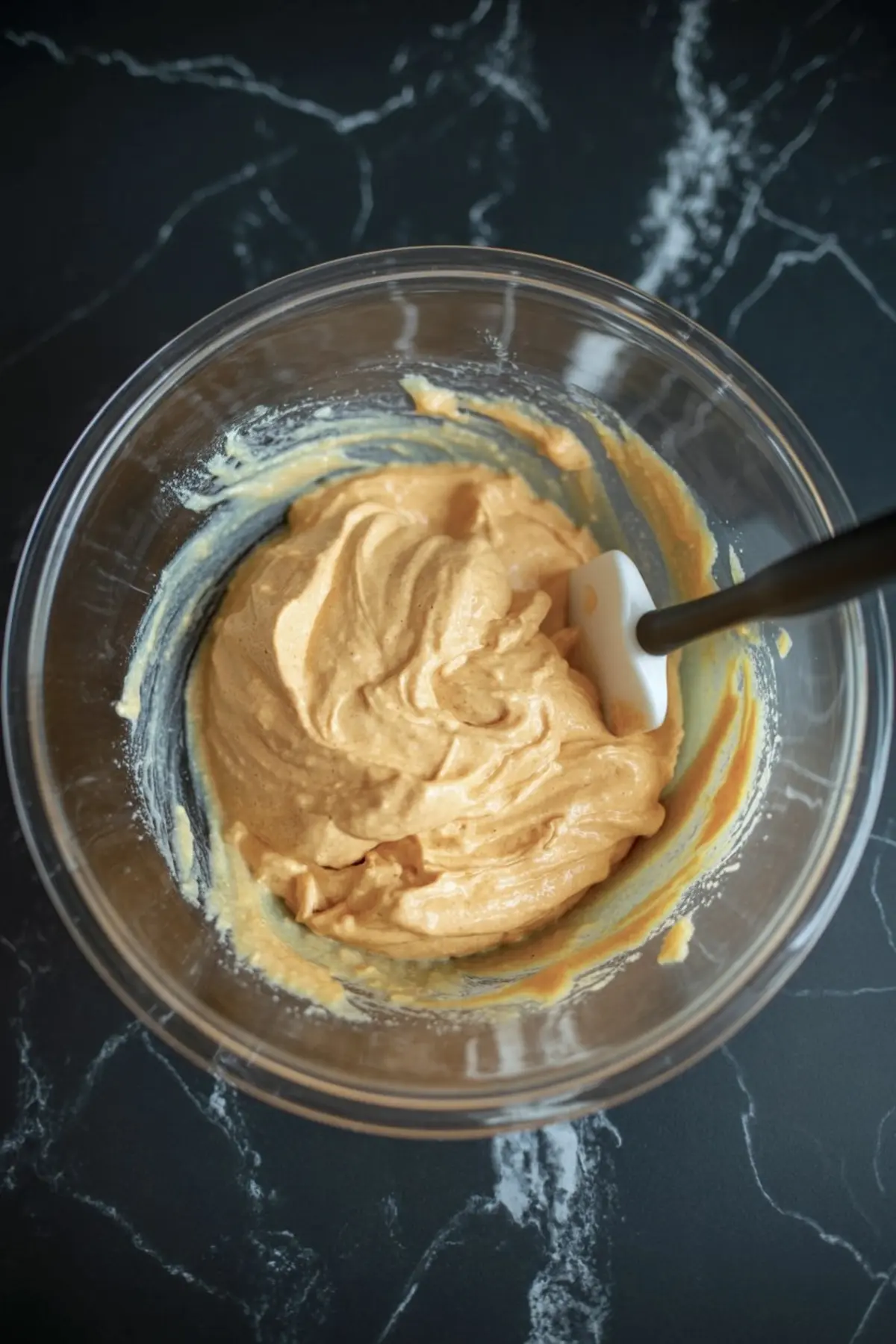 Creamy pumpkin cheesecake mixture in a clear mixing bowl with a spatula on a black marble surface.