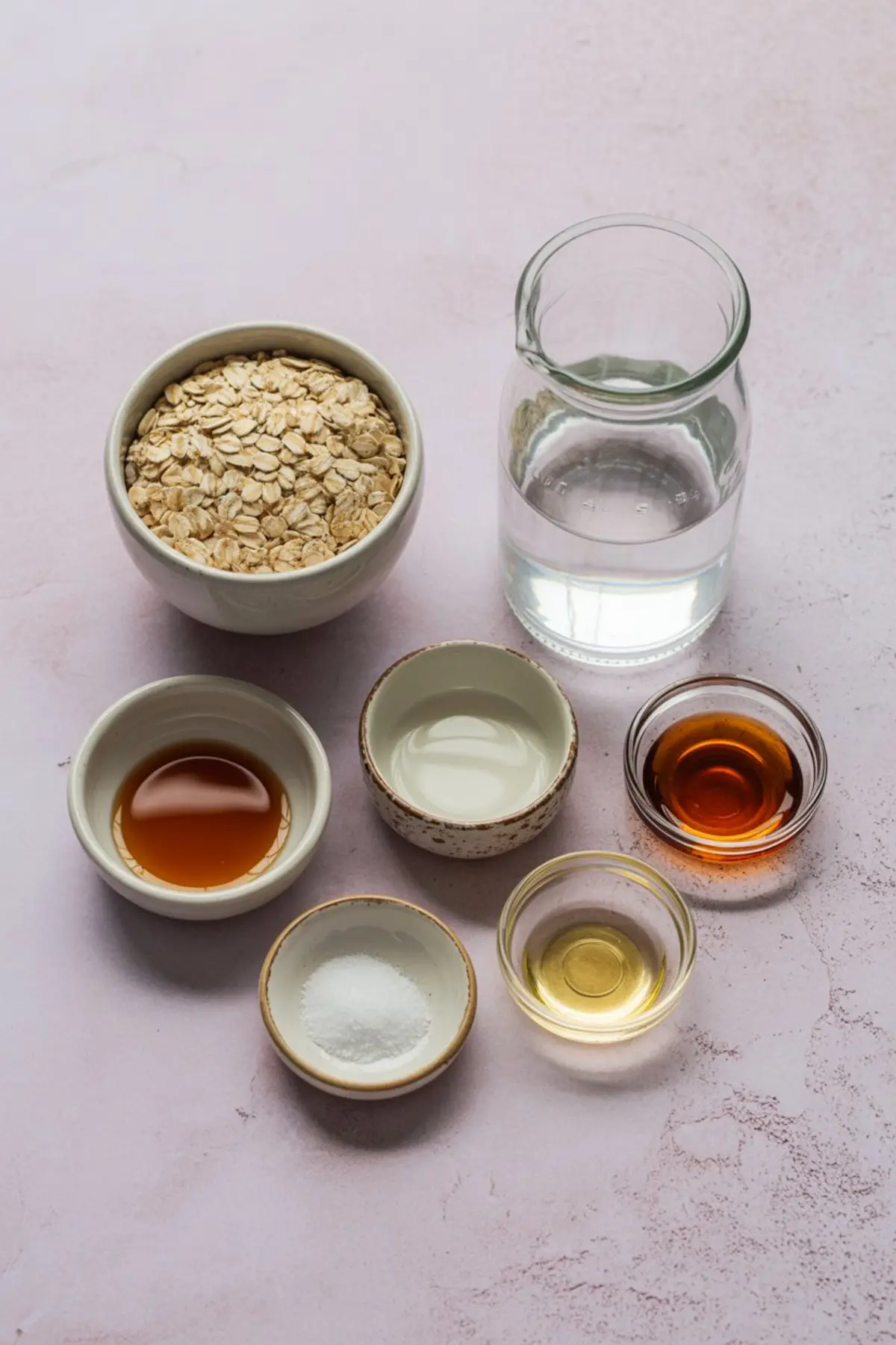 Flat lay of oat milk ingredients including rolled oats, water, maple syrup, vanilla extract, oil, and salt in small bowls on a pink background.
