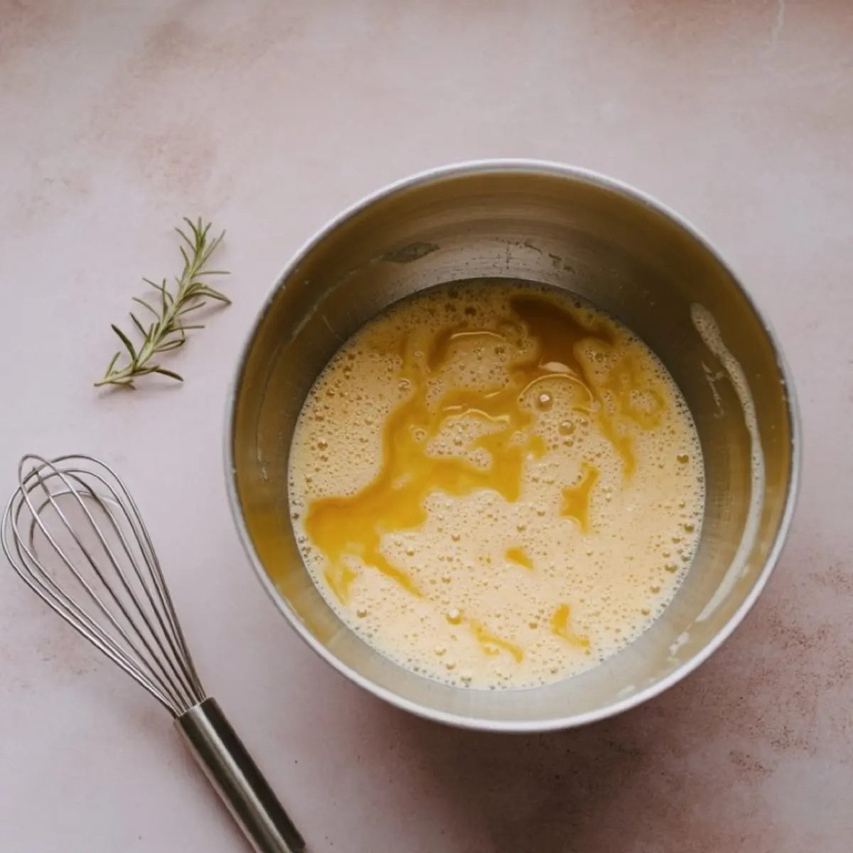 Stainless steel mixing bowl filled with beaten eggs and melted butter, placed on a light pink surface next to a whisk and a sprig of fresh rosemary.