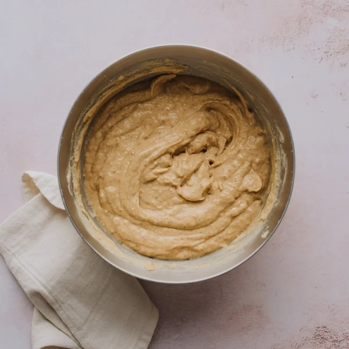 Thick pancake batter in a metal mixing bowl resting on a light pink counter with a soft cream kitchen towel beside it.
