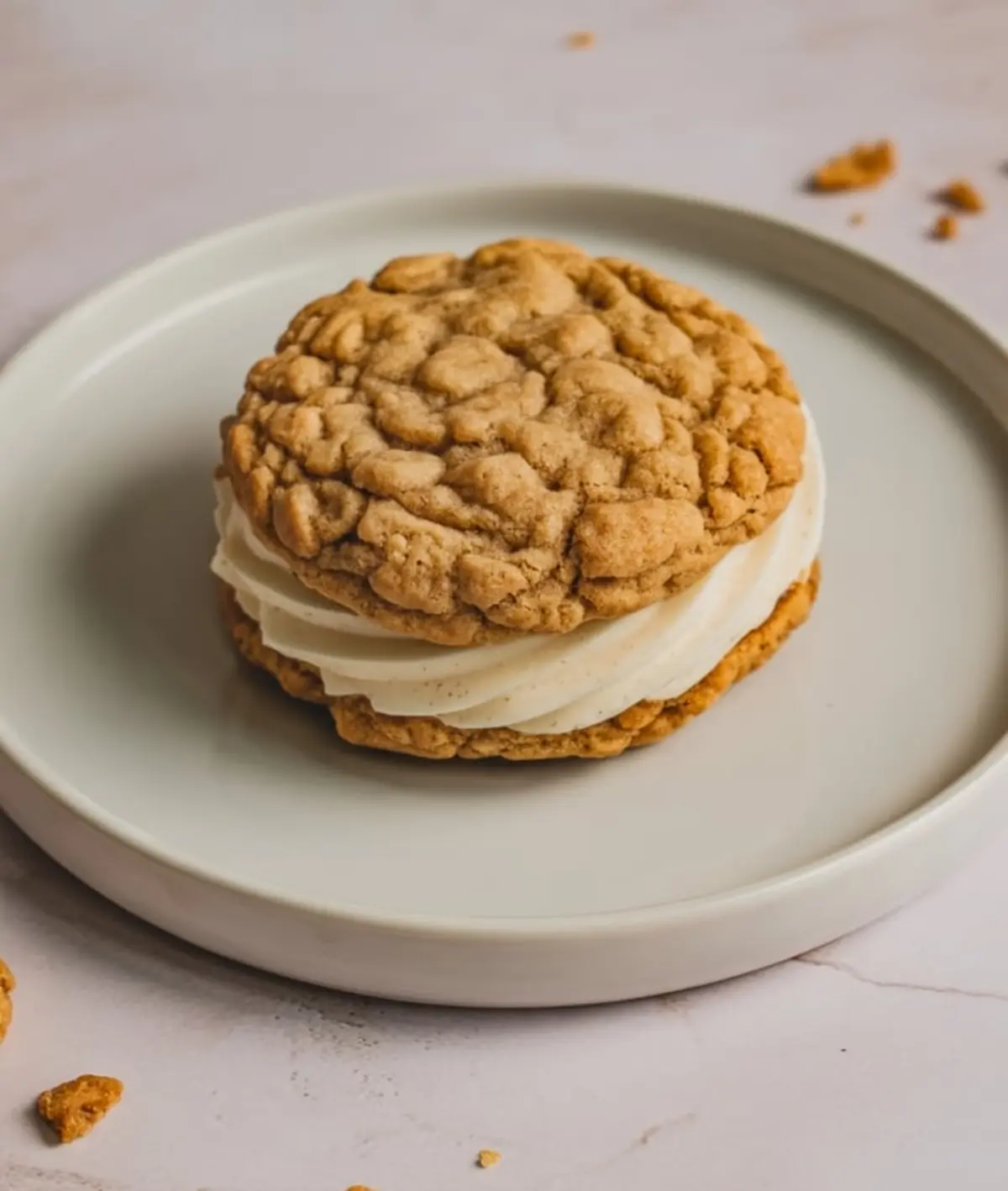 Oatmeal cream pie cookie with thick vanilla frosting filling, placed on a round white ceramic plate over a light marble background.