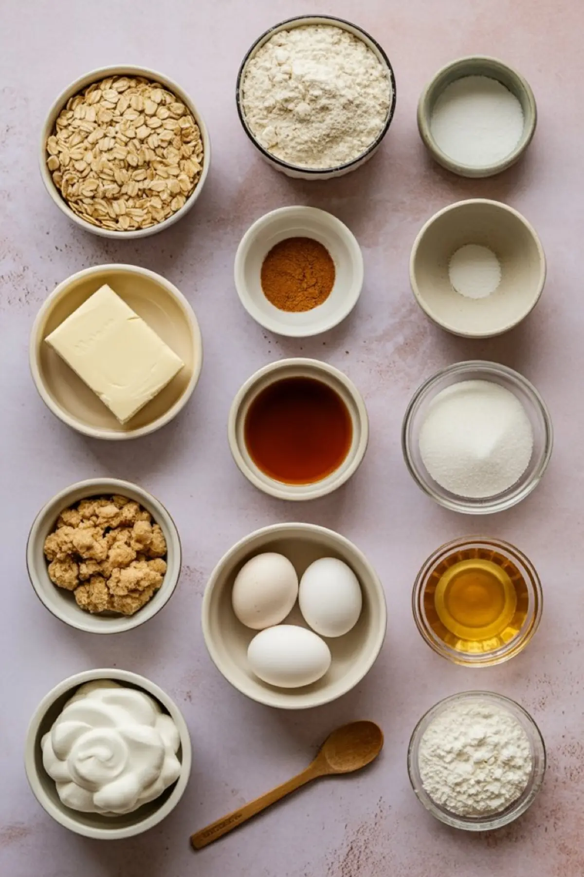 Flat lay of oatmeal cream pie cookie ingredients including rolled oats, flour, butter, eggs, brown sugar, granulated sugar, cinnamon, vanilla extract, baking soda, baking powder, and whipped frosting in small bowls on a pink background.