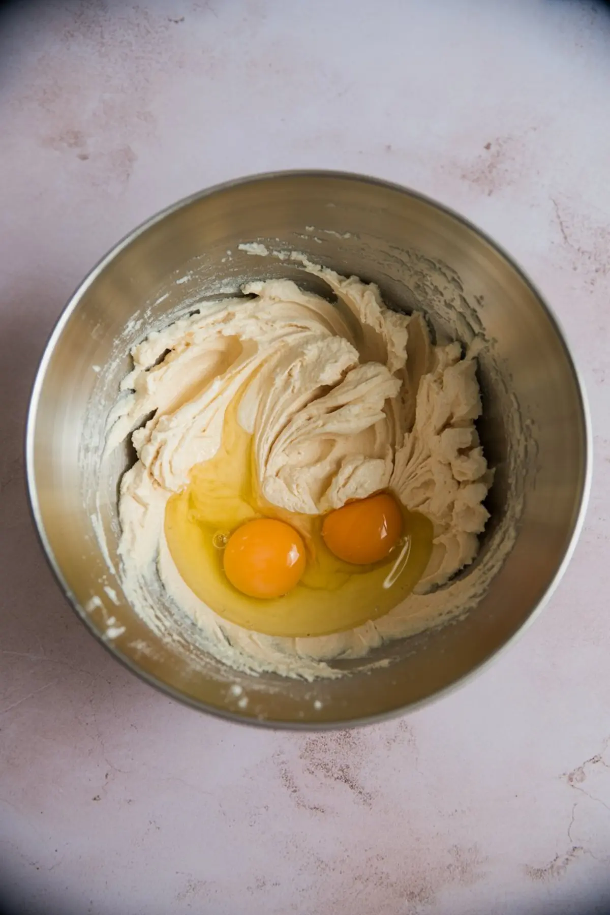 Metal mixing bowl with whipped butter and sugar mixture, topped with two cracked raw eggs on a pink countertop.