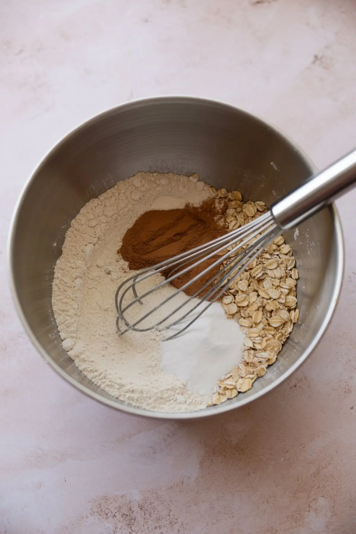 Dry ingredients for oatmeal cookies including oats, flour, baking soda, baking powder, and ground cinnamon in a mixing bowl with a whisk on the side.