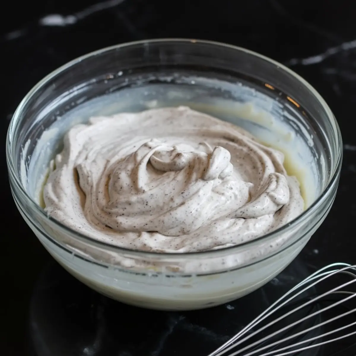 A glass bowl containing a fluffy Oreo whipped cream mixture with specks of crushed cookies, prepared for layering into no-bake desserts or Oreo cream pies.