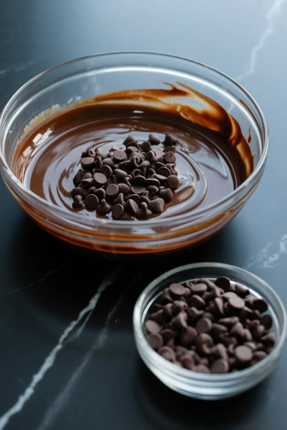 A clear bowl of melted chocolate with dark chocolate chips melting on top, set beside an extra bowl of chocolate chips on a black marble surface.