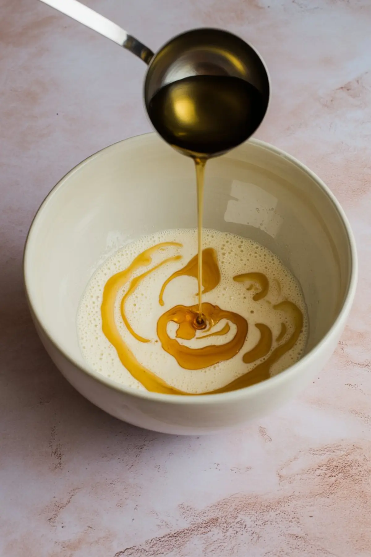 Golden syrup being poured into a bowl of foamy cream mixture, creating a swirl pattern against a pink textured surface.
