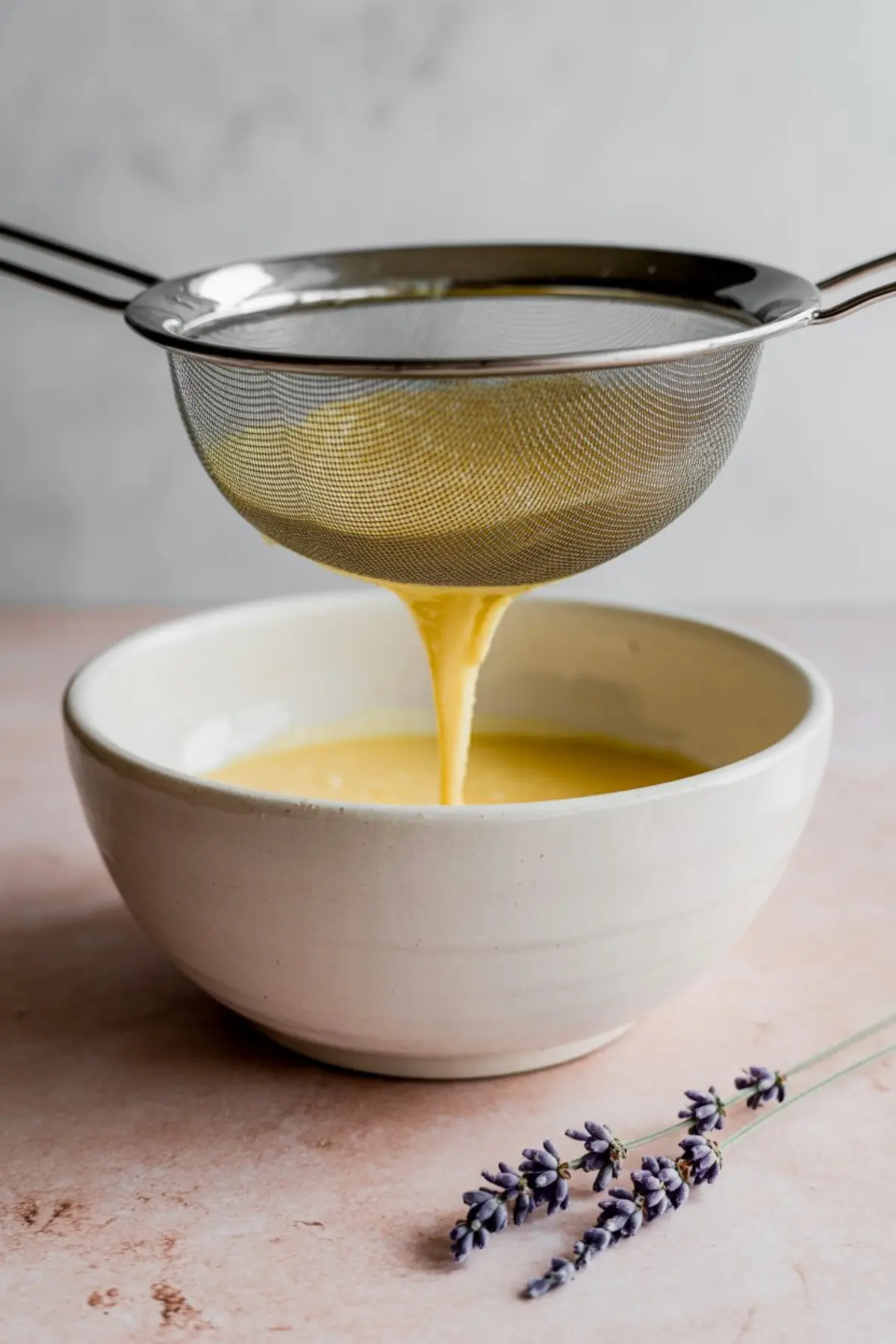 Smooth yellow custard being strained through a fine mesh sieve into a white bowl, with sprigs of lavender resting nearby.
