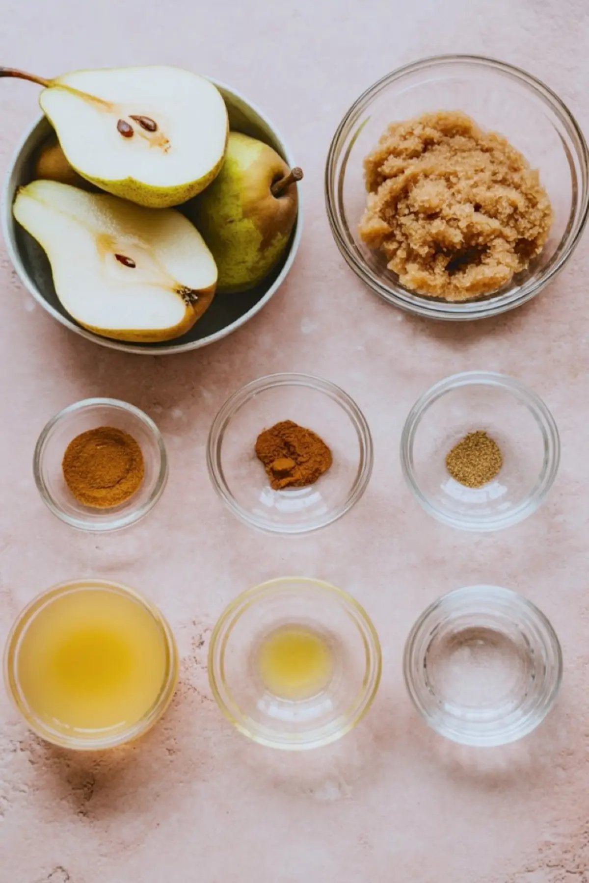 A flat lay of ingredients for pear butter, including fresh pears, brown sugar, ground spices, apple juice, lemon juice, and water arranged in glass bowls on a light pink background.