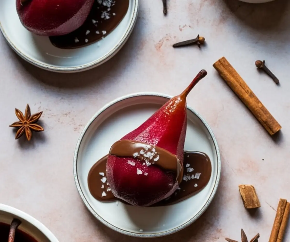 Poached pear on a white plate topped with creamy chocolate sauce and sea salt, surrounded by cinnamon sticks, star anise, and cloves on a textured pink surface.