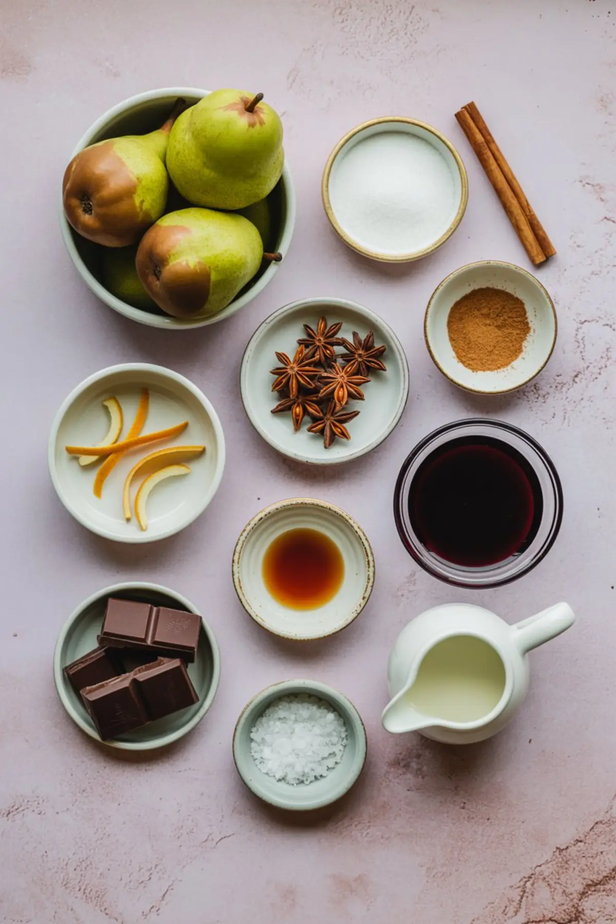 Overhead view of poached pear ingredients including fresh green pears, sugar, cinnamon sticks, orange peel, star anise, red wine, vanilla, milk, sea salt, and chocolate, arranged in small bowls on a light pink background.