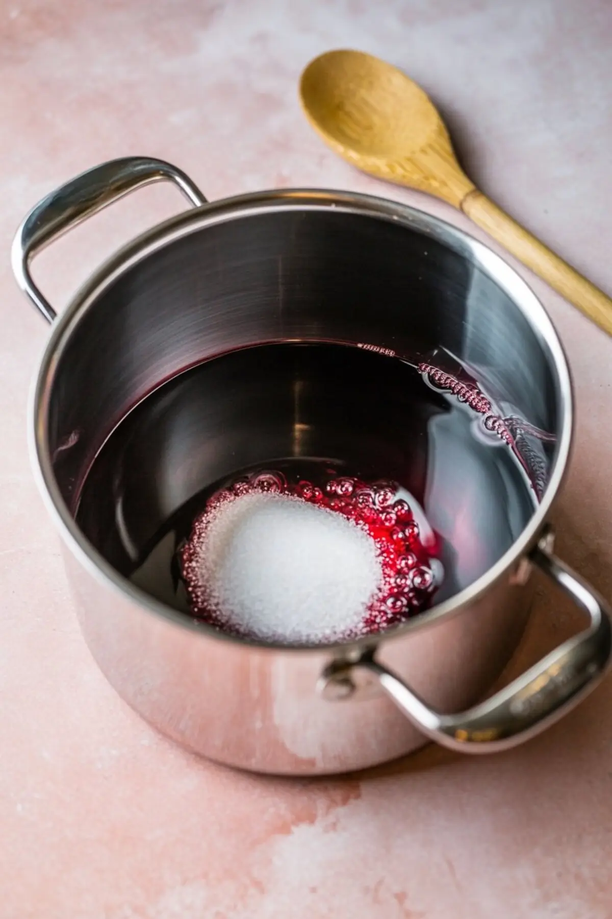 Sugar being added to red wine in a stainless steel pot, placed on a light pink surface with a wooden spoon beside it, beginning the poaching syrup preparation.