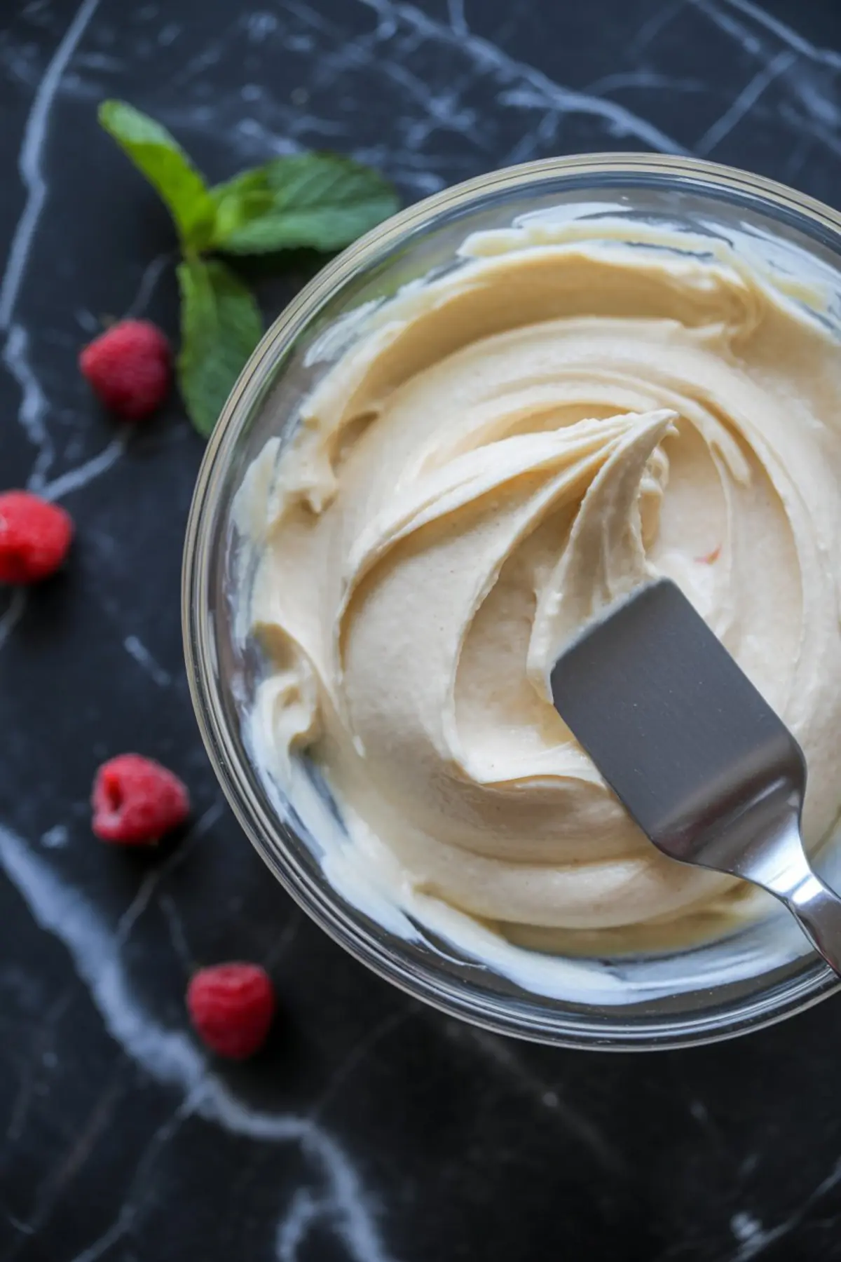 Creamy frosting being spread in a glass bowl with a metal spatula, surrounded by fresh raspberries and mint leaves.