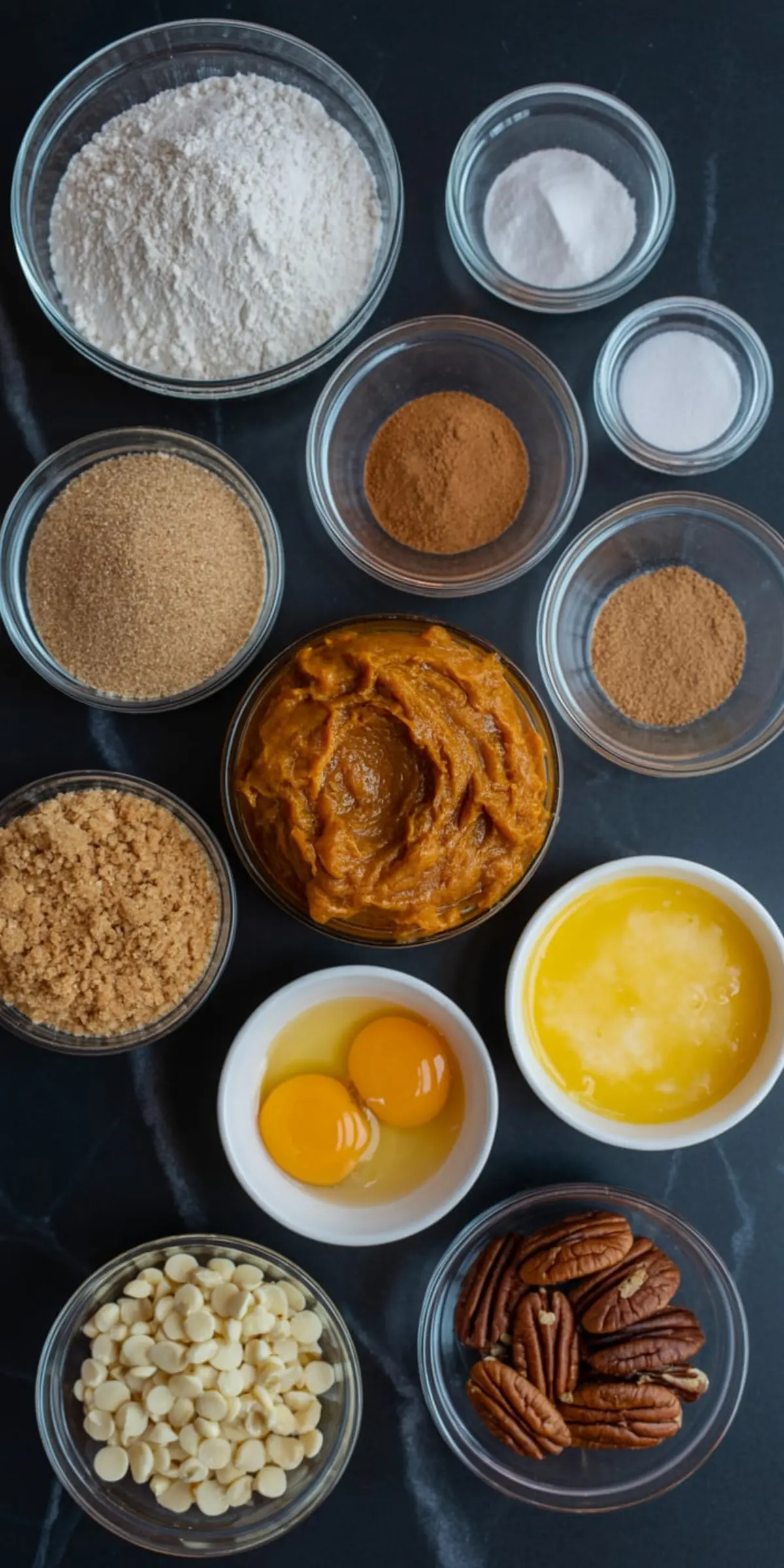 Overhead view of pumpkin cookie ingredients on a dark countertop, including flour, baking soda, salt, brown sugar, pumpkin puree, eggs, melted butter, white chocolate chips, pecans, and warm spices like cinnamon and nutmeg.