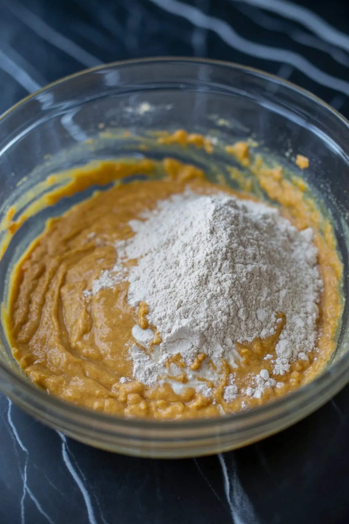 Flour added to thick pumpkin cookie dough inside a clear mixing bowl, midway through combining dry and wet ingredients.