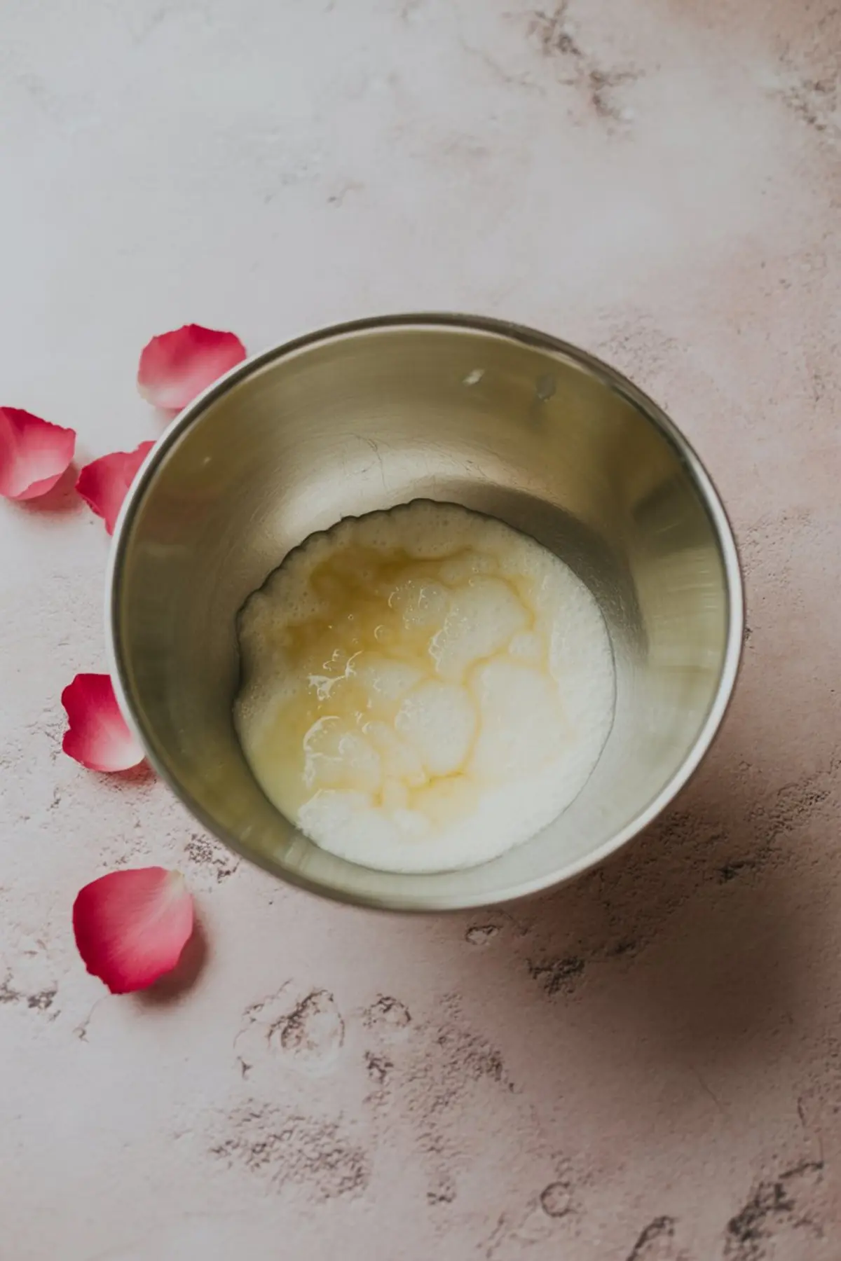 Metal mixing bowl containing whipped egg whites and sugar on a pink background, with rose petals around the edges, showing the preparation of a fluffy pie filling.
