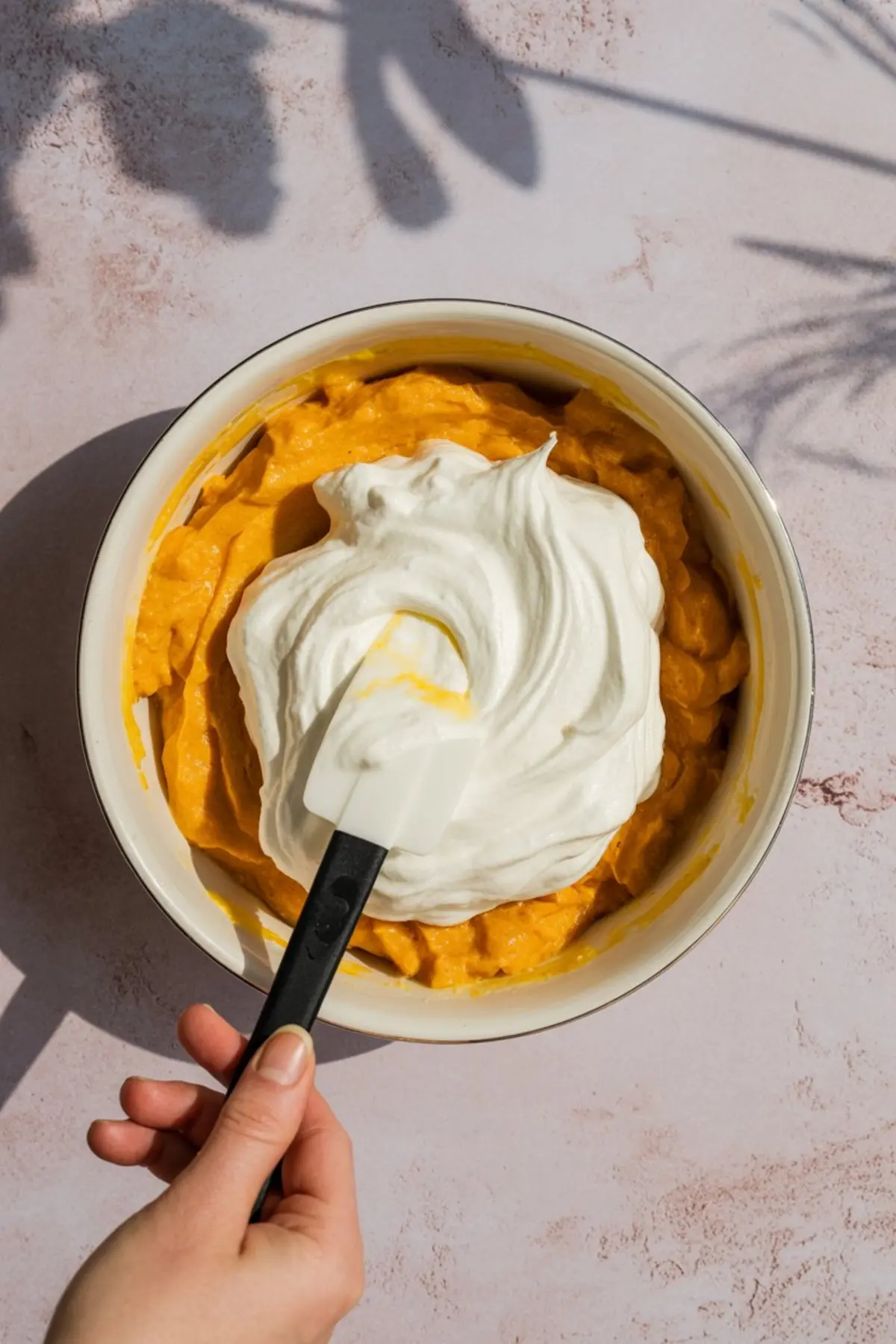 Hand holding a spatula folding whipped cream into a bowl of vibrant orange pumpkin mixture, preparing a light and airy chiffon pie filling on a soft pink background with natural shadows.