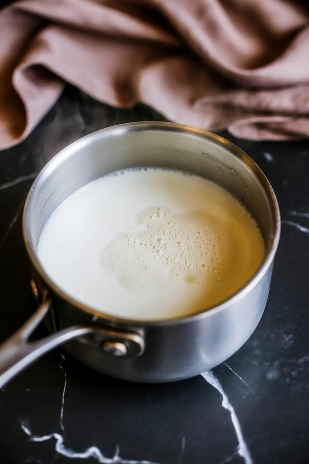 Stainless steel saucepan filled with warm milk on a black marble countertop with a soft beige cloth in the background.