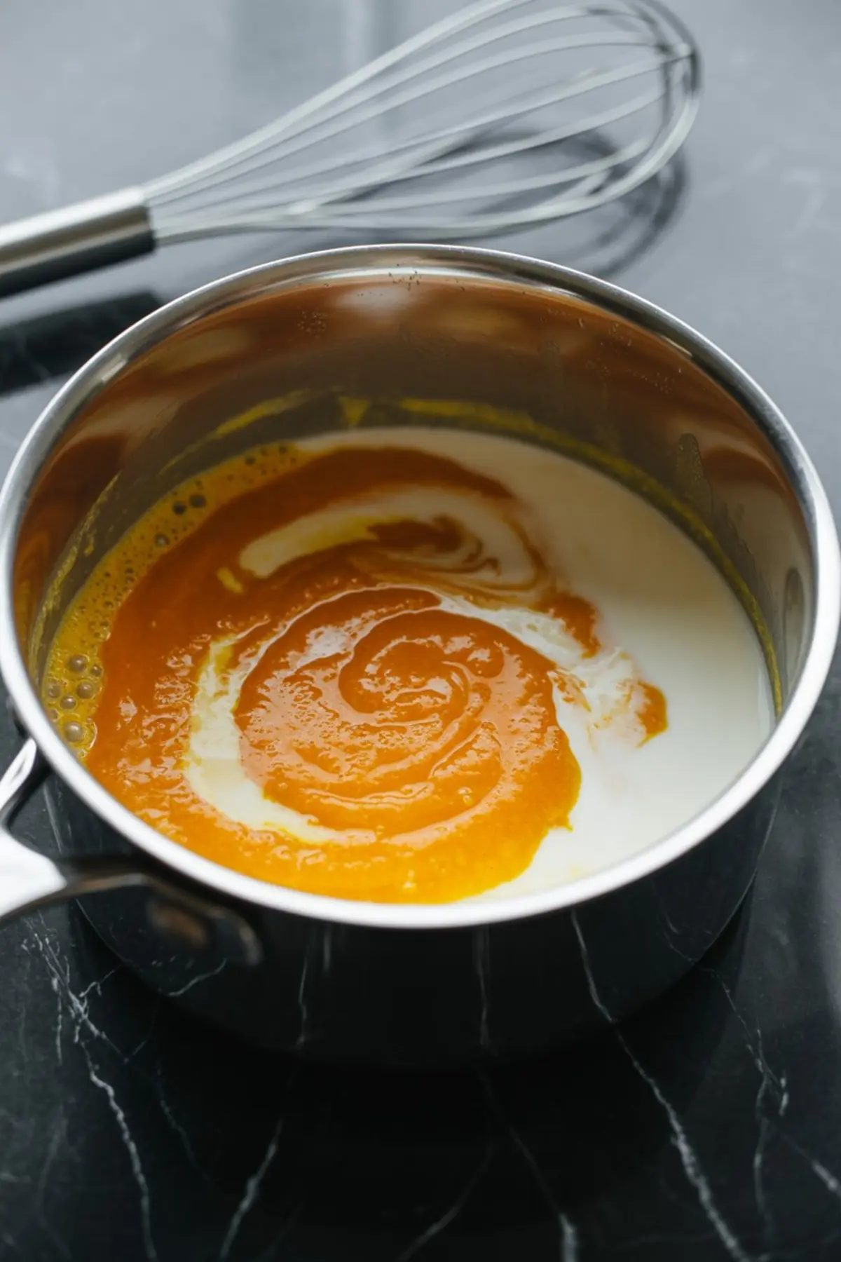 Saucepan on a dark countertop with pumpkin puree being swirled into warm milk, with a metal whisk nearby.