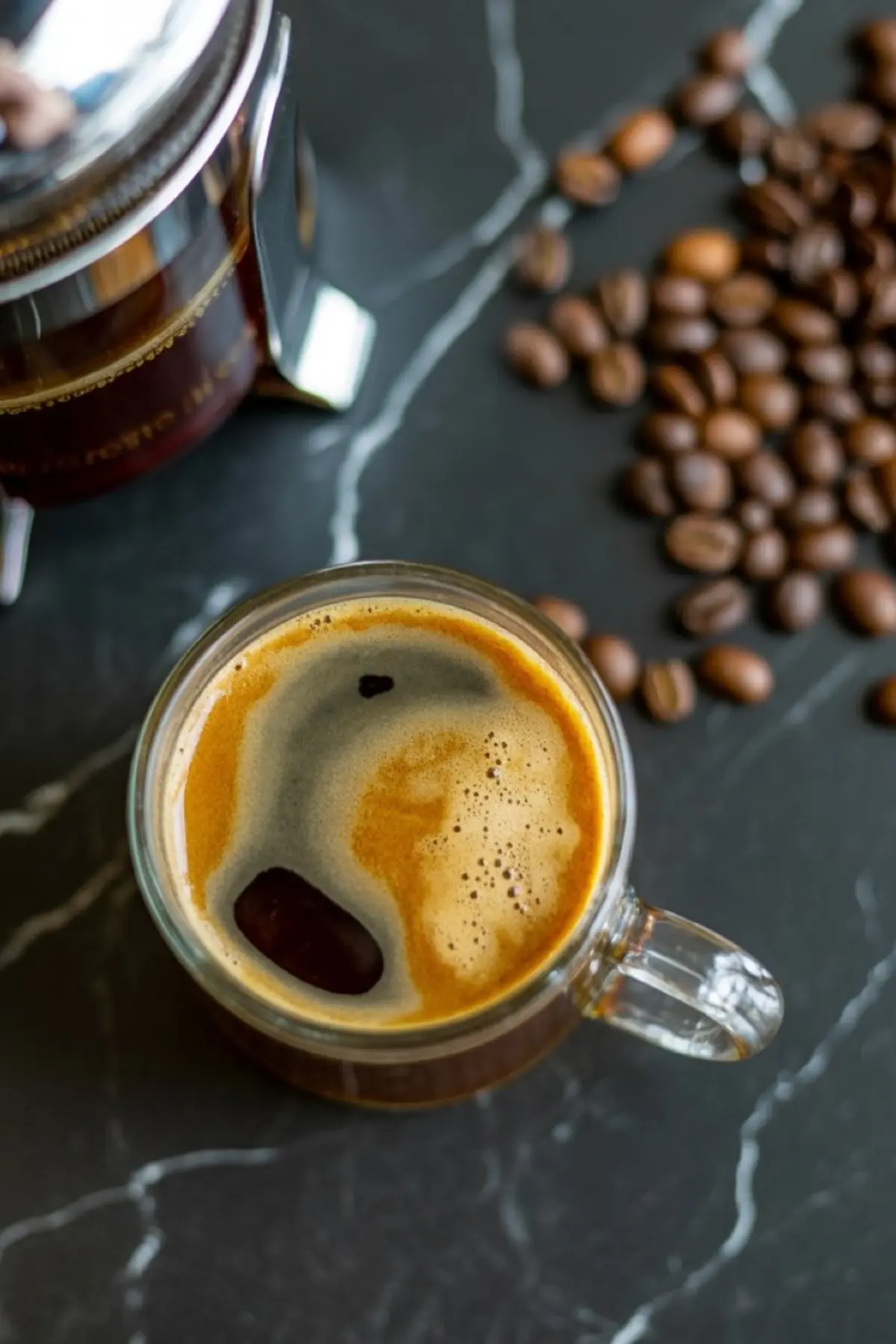 Glass mug of freshly brewed black coffee with creamy crema on top, placed beside whole coffee beans and a French press on a black marble surface.
