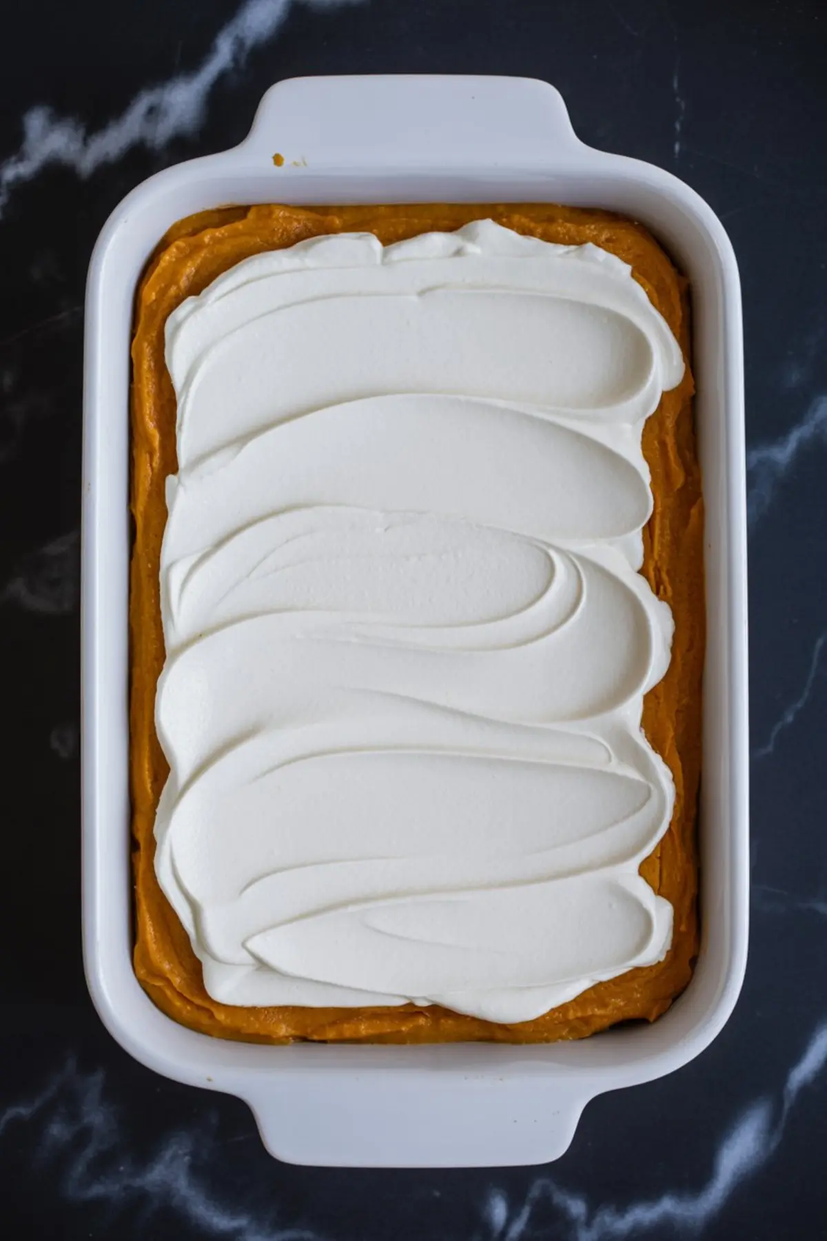 Pumpkin cream cheese dessert in a white baking dish, topped with a thick layer of whipped cream swirled in smooth, even strokes.