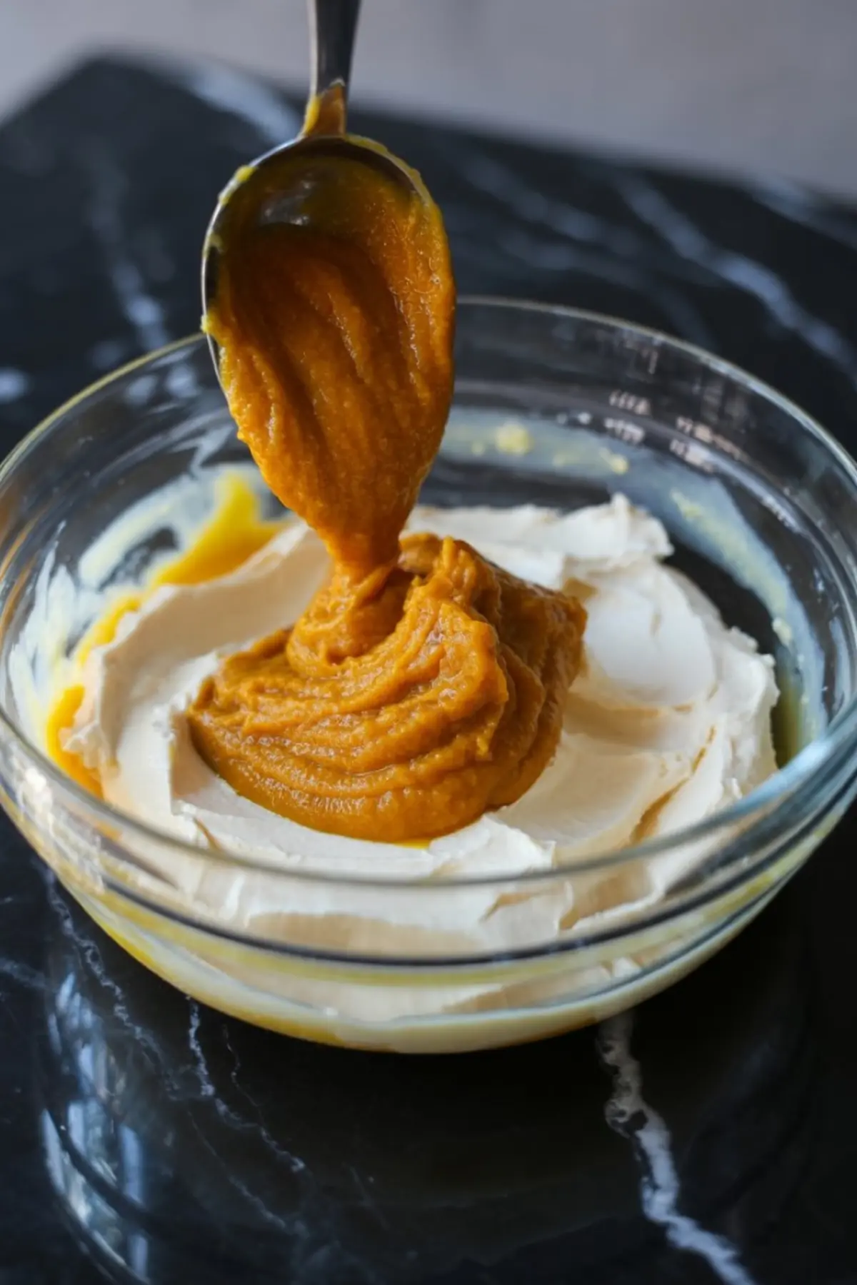 Spoonful of pumpkin purée being added to a bowl of whipped cream cheese, showing the process of blending ingredients for pumpkin cream cheese. This preparation scene captures seasonal recipe creation.
