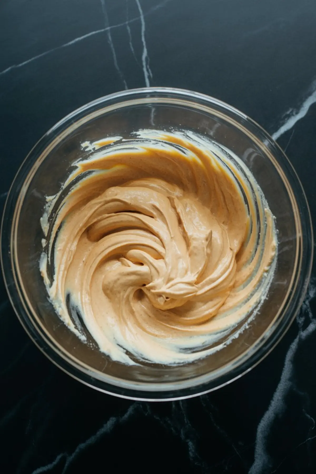 Glass mixing bowl with freshly blended pumpkin cream cheese, showing a smooth and creamy orange-tinted swirl on a dark marble surface. The texture captures the rich consistency of the autumn spread.
