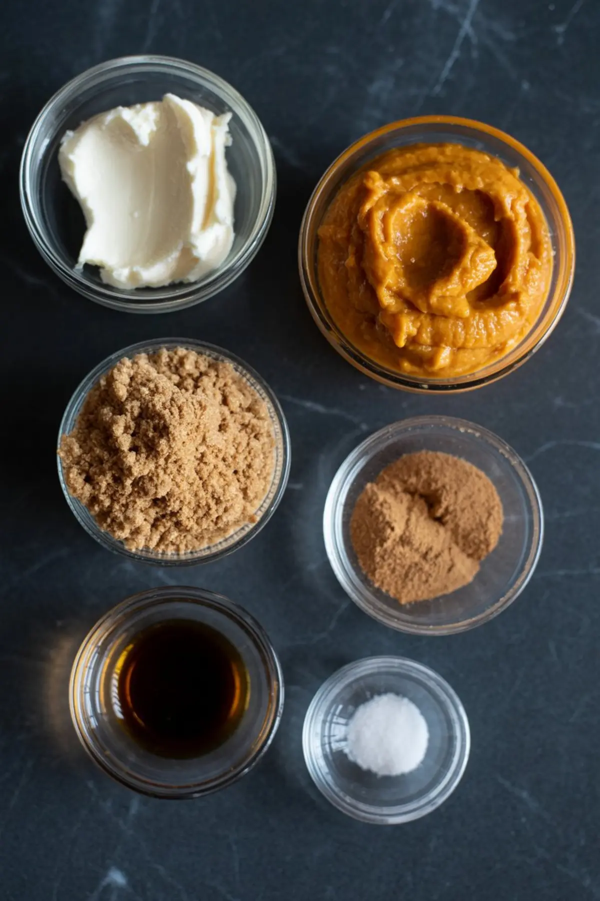 Flat lay of ingredients for pumpkin cream cheese on a dark surface, including cream cheese, pumpkin purée, brown sugar, cinnamon, vanilla extract, and salt. This setup visually highlights the components for a homemade fall-flavored spread.
