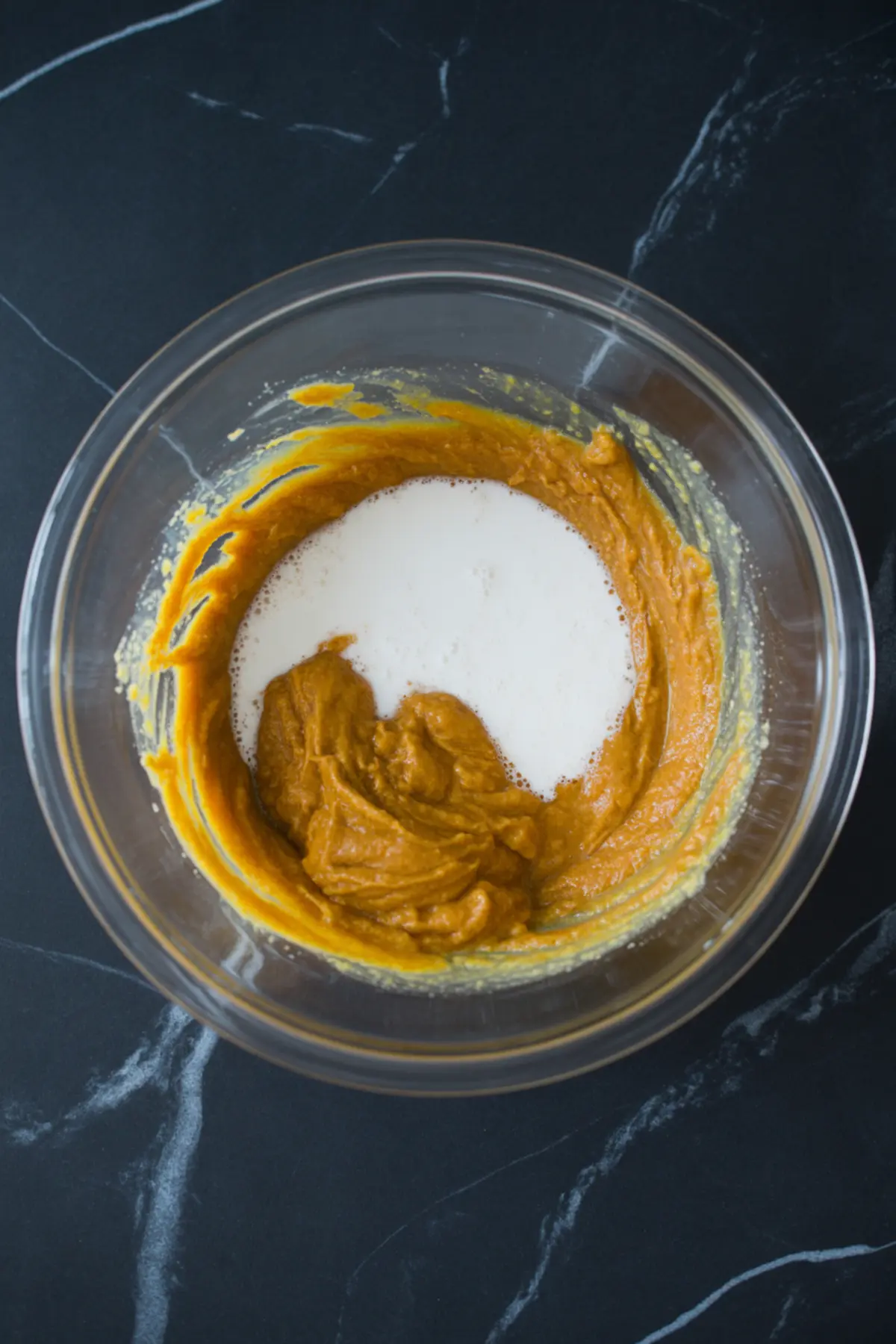Glass mixing bowl filled with pumpkin puree and cream being combined for a custard base on a black marble surface.