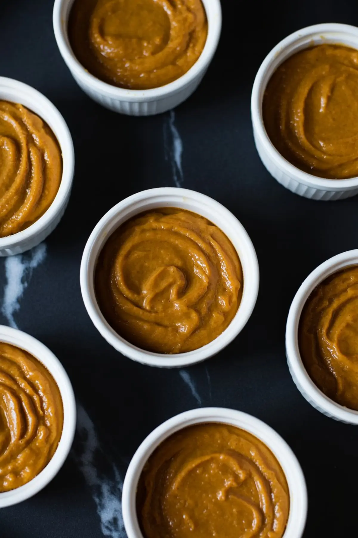 White ramekins filled with smooth pumpkin custard mixture before baking, displayed on a black marble background.