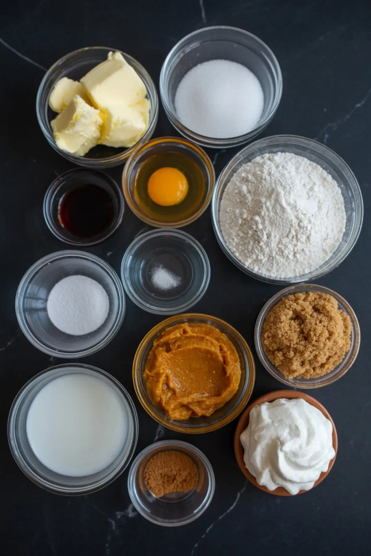 Flat lay of individual baking ingredients in glass bowls including butter, egg, sugar, flour, pumpkin purée, brown sugar, milk, vanilla extract, baking soda, spices, and whipped cream on a dark countertop.