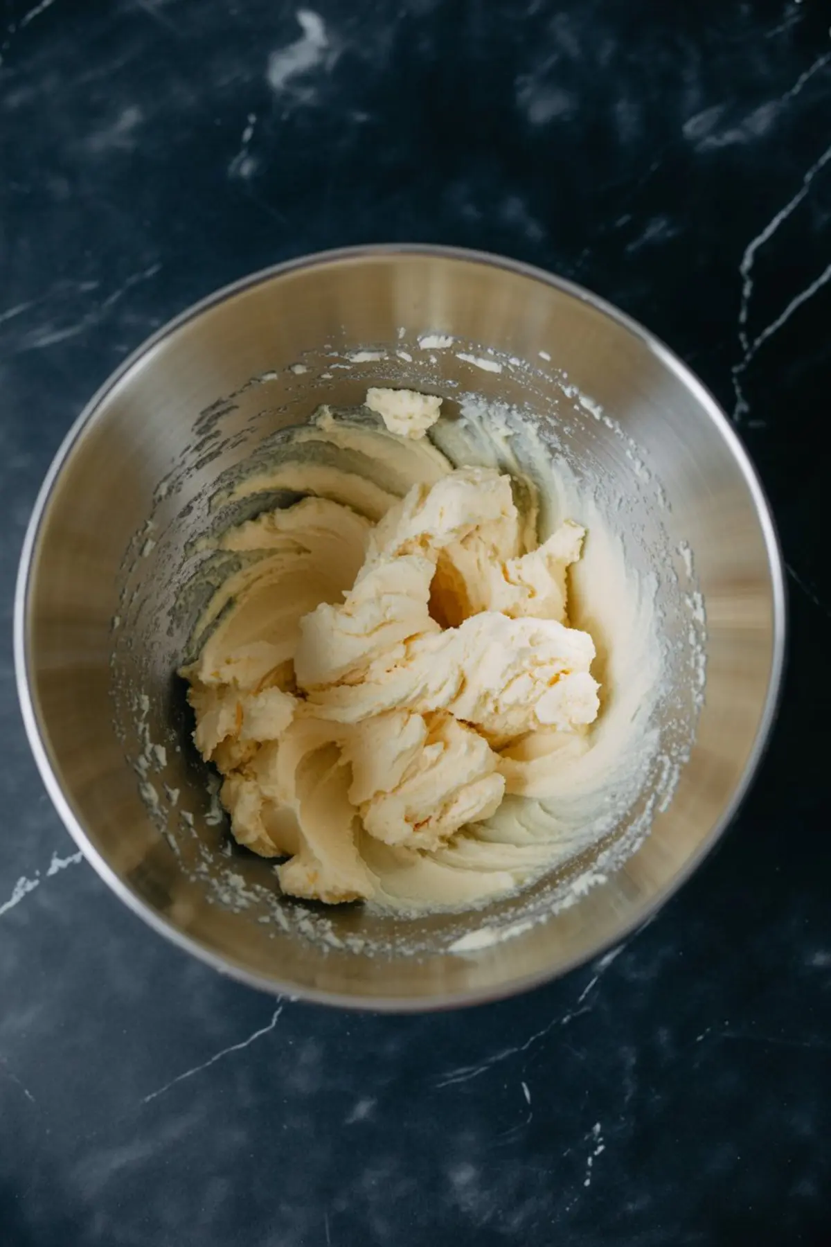 Creamed butter and sugar mixture in a stainless steel mixing bowl on a black marble counter, showing the base texture for cookie dough preparation.