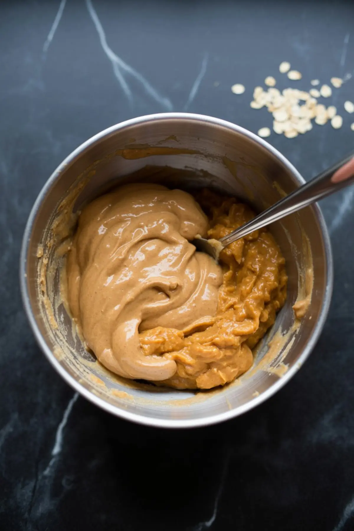 Thick pumpkin protein batter being combined with smooth peanut butter mixture in a metal bowl using a spoon, creating a creamy dough.
