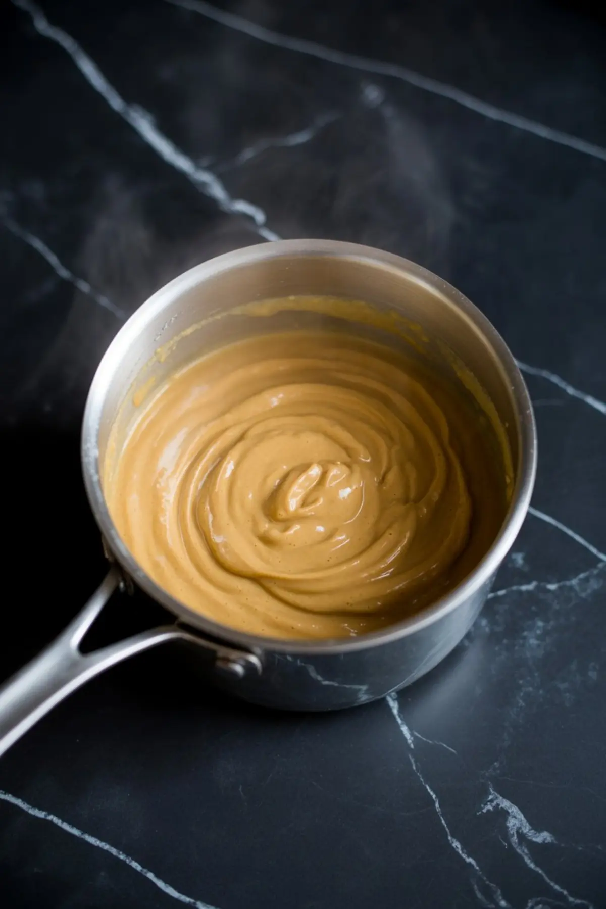 A saucepan filled with warm, thickened pumpkin pudding on a black marble counter. The creamy mixture is smooth and glossy, showing signs of being freshly cooked and stirred.