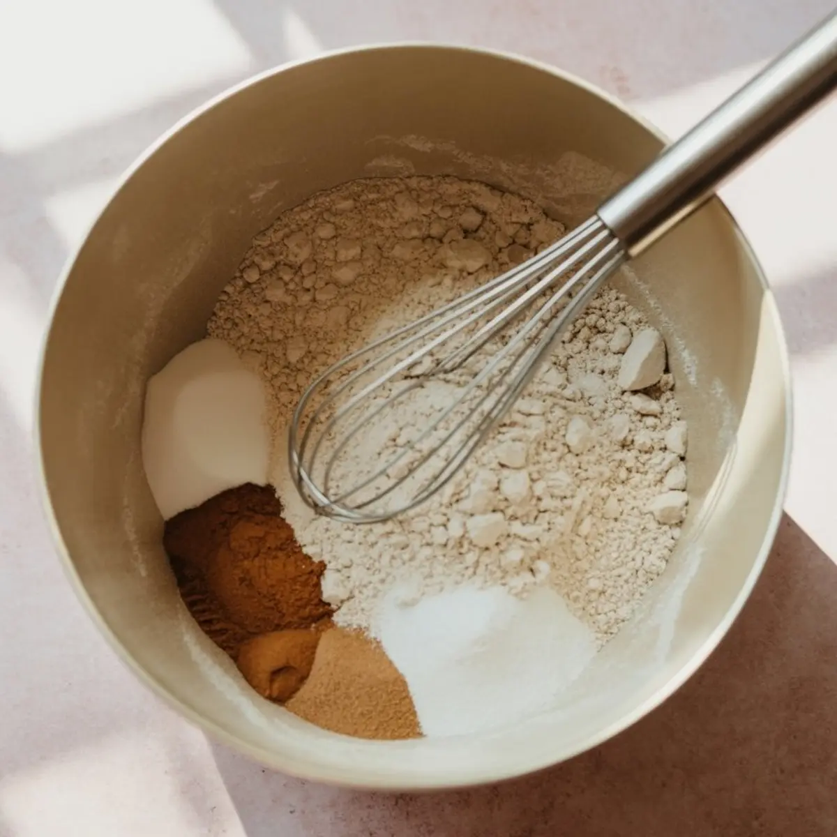 Mixing bowl with flour, sugar, cinnamon, nutmeg, and baking powder with a metal whisk, set on a sunlit pink surface for sweet potato pancake batter.
